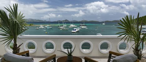 Un balcón con dos sillones de mimbre y una mesita, con vista a un puerto deportivo lleno de barcos y un mar azul cristalino.