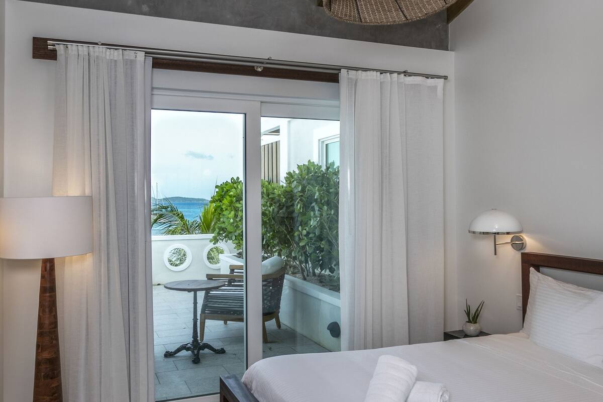 A bedroom with a wooden ceiling, a large bed with white linens, a floor lamp, and a view of the outdoors through a sliding glass door.