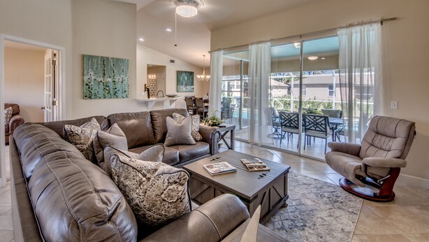 A spacious living room with a leather sofa, a recliner, and a coffee table. There is a large window with curtains, a dining area in the background, and a view of the outdoors. - 2621 SW 27th St. Cape Coral (Cape Coral)