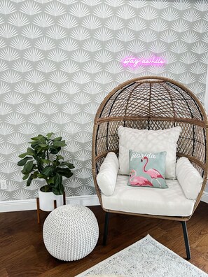 Una silla de mimbre con un cojín y una almohada, una planta en maceta y un letrero de neón en una habitación con papel tapiz estampado.