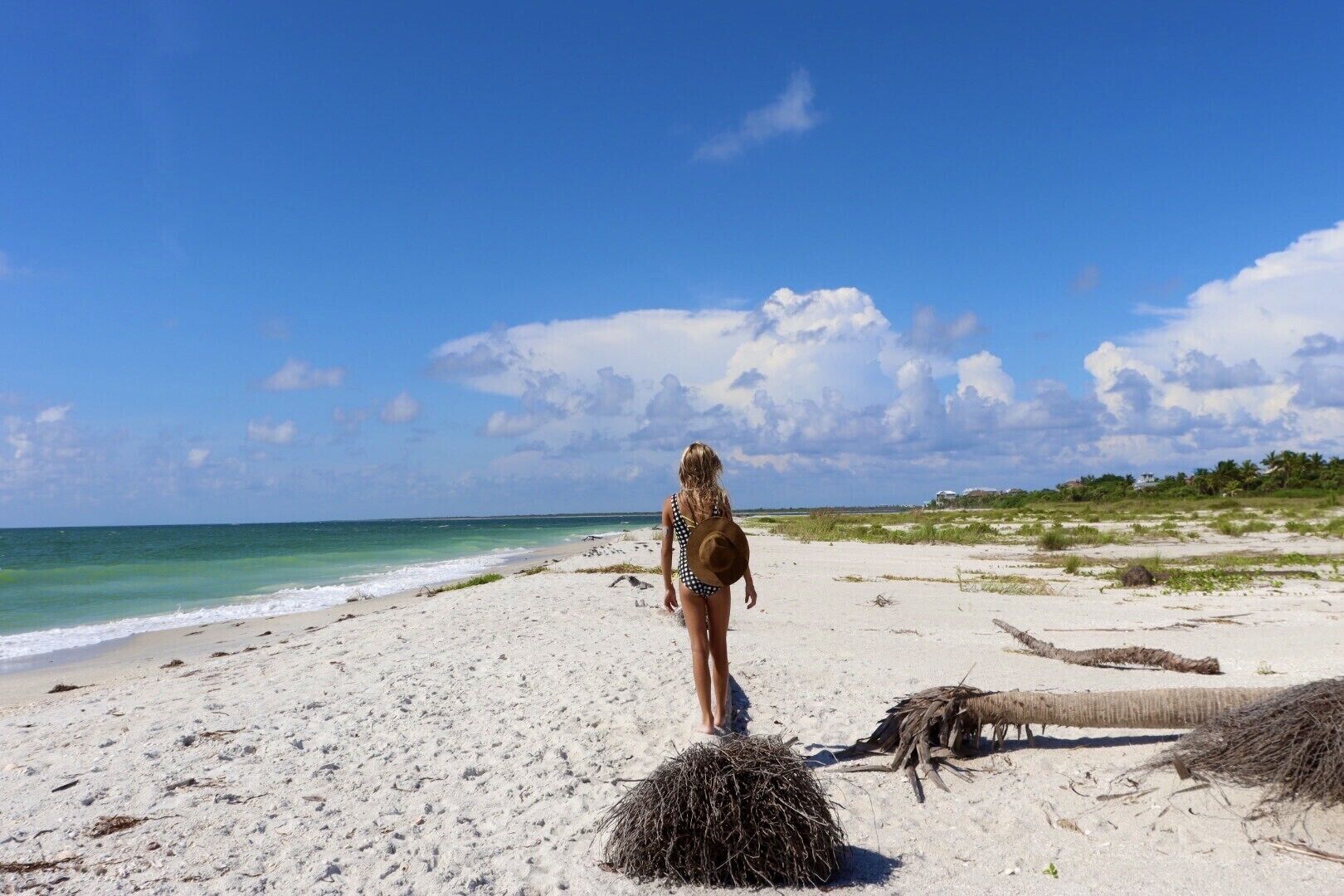En kvinde i badetøj går på en sandstrand med hav og vegetation i baggrunden.
