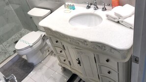 Une salle de bain avec un lavabo, un meuble-lavabo et des toilettes, tous de couleur blanche. On aperçoit des articles de toilette sur le lavabo et un rideau de douche à l’arrière-plan.