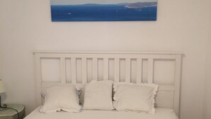 Un lit avec une tête de lit blanche et trois oreillers, une table de chevet avec une lampe, et un tableau représentant un paysage côtier accroché au mur.