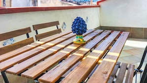 Una mesa de madera con bancos bajo un toldo a rayas, un jarrón decorativo y vistas de edificios y colinas al fondo.