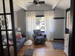 Un salon moderne avec un canapé gris, un fauteuil à motifs, une petite table ronde, un ventilateur de plafond et une vue sur la rue à travers une fenêtre avec des rideaux.