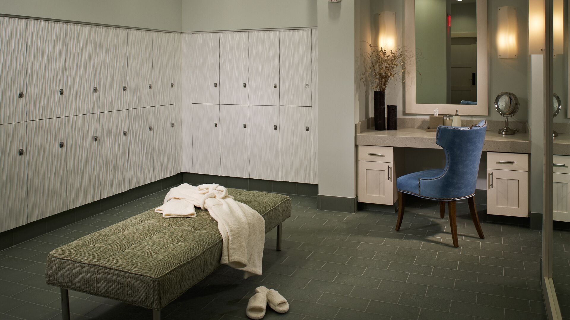 Un baño moderno con un banco, una silla azul, un tocador con espejo y una pared de taquillas.