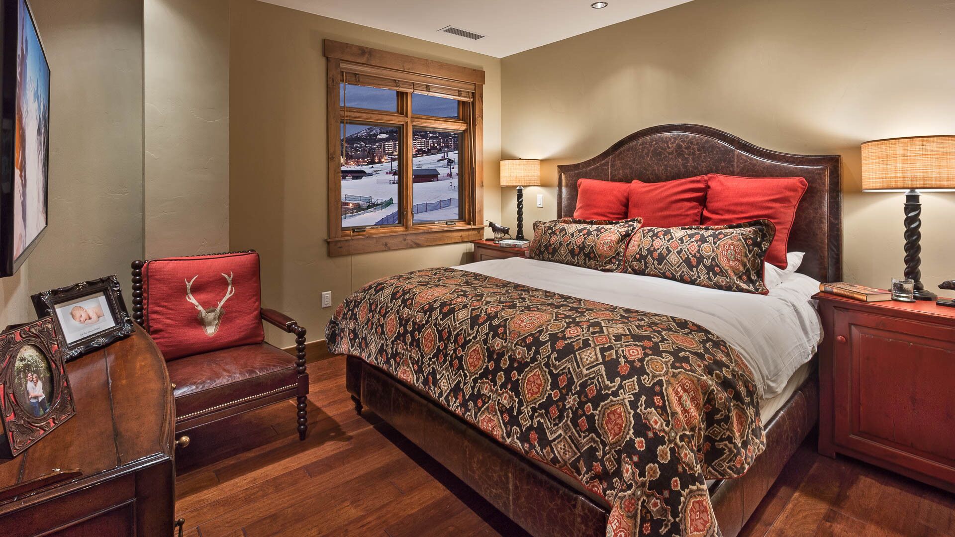 Une chambre à coucher avec un grand lit, une tête de lit en bois, une table de chevet, une chaise avec un coussin rouge, un tableau encadré et une fenêtre donnant sur un paysage enneigé.