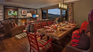 A dining area with a wooden table set for six, surrounded by leather chairs. A fireplace and a television are visible in the background.