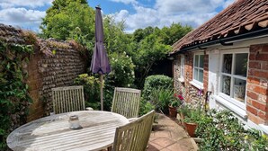 A rustic outdoor seating area with a round wooden table and two chairs, a purple umbrella, and a stone wall.