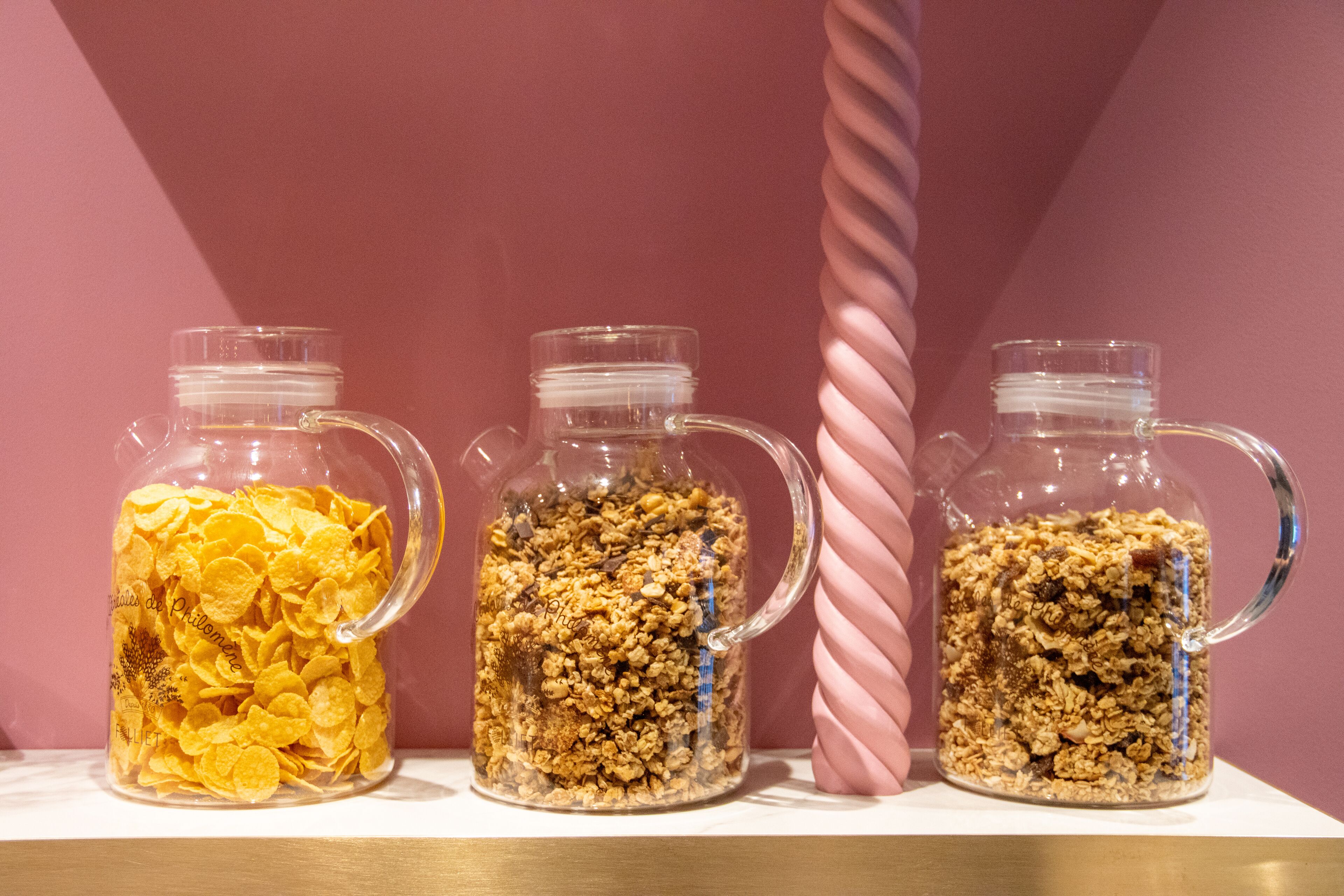 Three glass jars filled with different types of cereal on a shelf against a pink background.