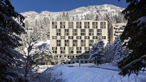 A multi-story building with a symmetrical facade, surrounded by snow-covered trees and a mountainous backdrop.