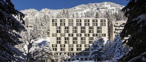 Un edificio a più piani con una facciata simmetrica, circondato da alberi innevati e uno sfondo montuoso.
