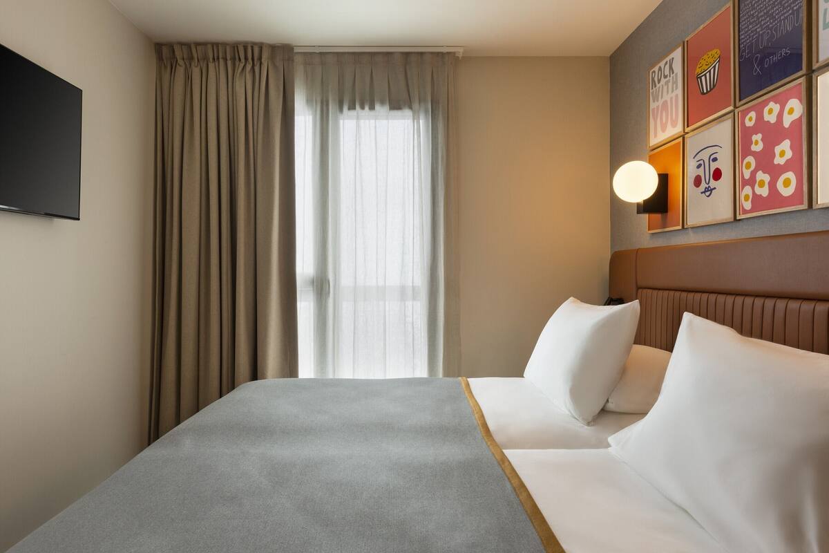 Een hotelkamer met een bed, een raam met gordijnen, een wandtelevisie en een muur met ingekaderde foto's.