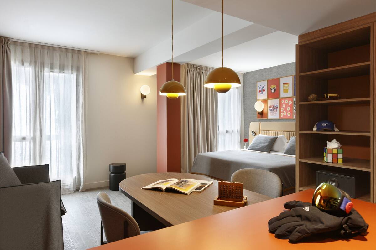 Hotelkamer met een bed, een bureau met een boek, een stoel, een tafel, een plank met spullen en een raam met gordijnen.