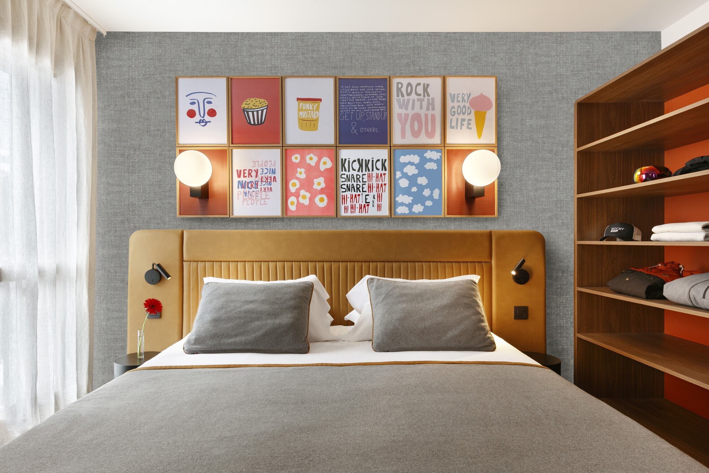 A neatly arranged hotel room with a large bed, a headboard, a wall-mounted shelf with neatly folded clothes, and a series of framed motivational posters.
