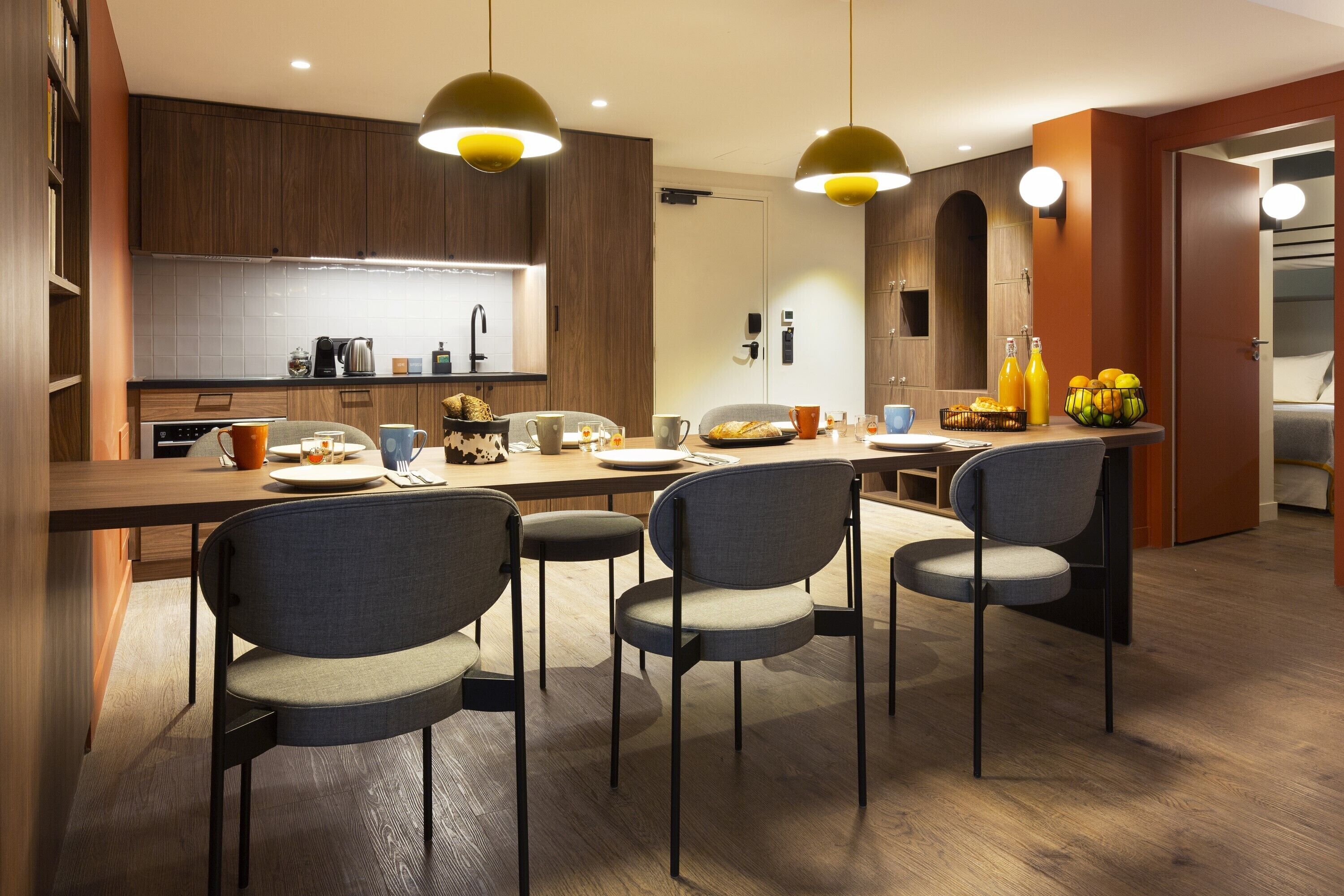 Eine moderne Küche mit Holzausstattung, einer zentralen Kochinsel mit Stühlen, Pendelleuchten und einer Frühstücksecke mit Früchten und Getränken.