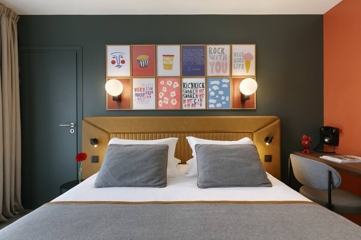 Een moderne hotelkamer met een groot bed, twee kussens, een houten hoofdbord, een bureau met stoel, en een wand met kleurrijke kunstwerken.
