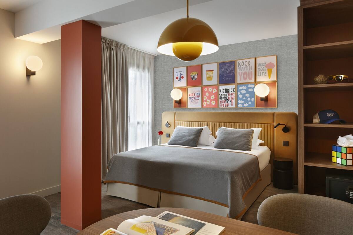 Een hotelkamer met een bed, nachtkastje, lamp, een boekenplank en een raam met gordijnen.