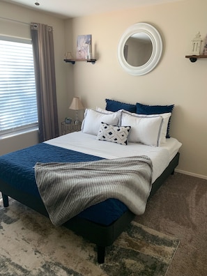Une chambre à coucher comprenant un lit, une table de chevet, une lampe, une fenêtre avec des rideaux, un miroir et une étagère fixée au mur.
