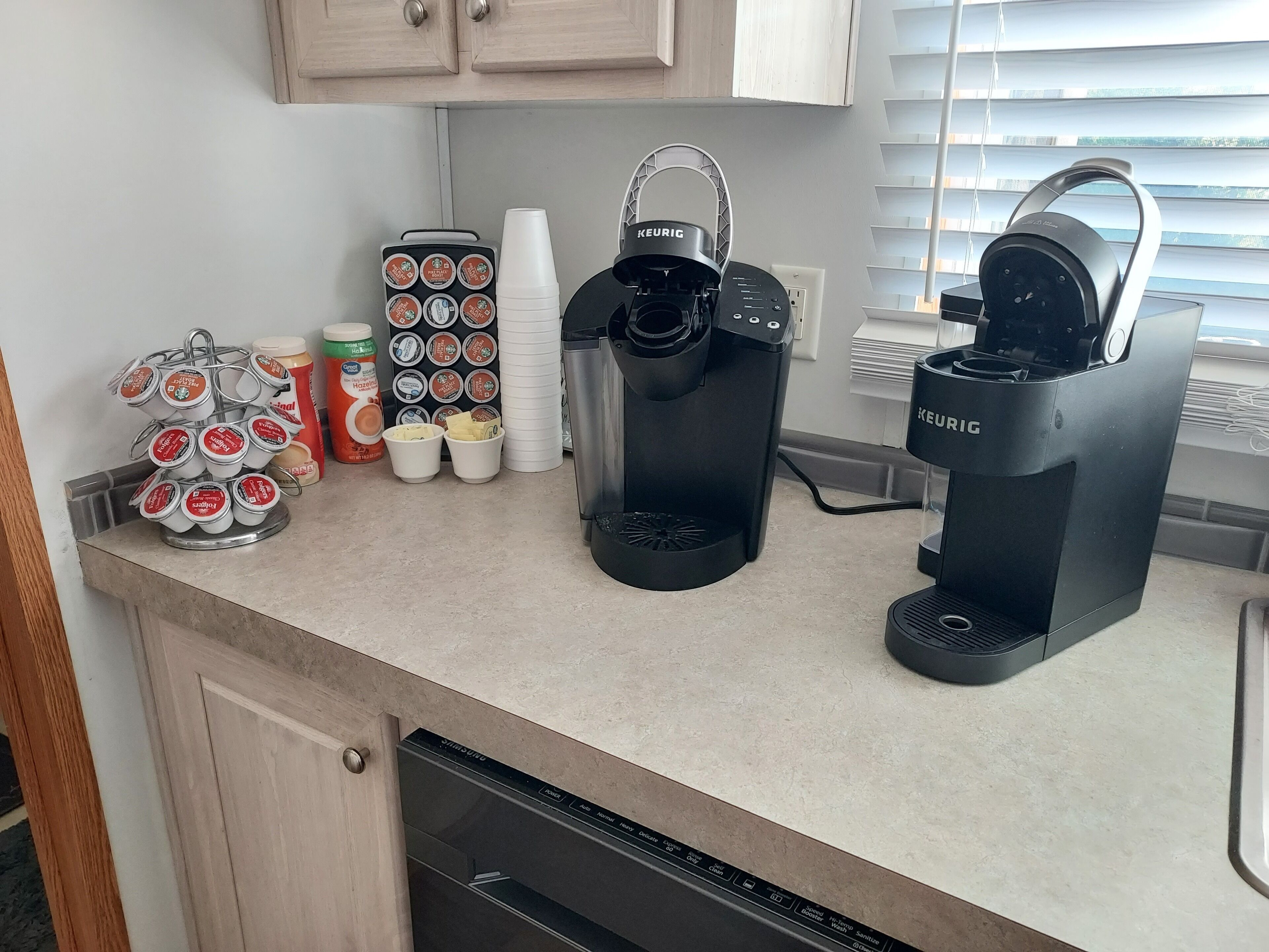 A kitchen counter with a Keurig coffee maker, a Keurig water dispenser, a coffee machine, a mug rack, and a coffee pod holder.