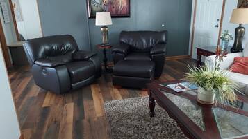A living room with two black leather chairs, a small round table with a lamp, a glass coffee table with a plant, and a white sofa with a red cushion.