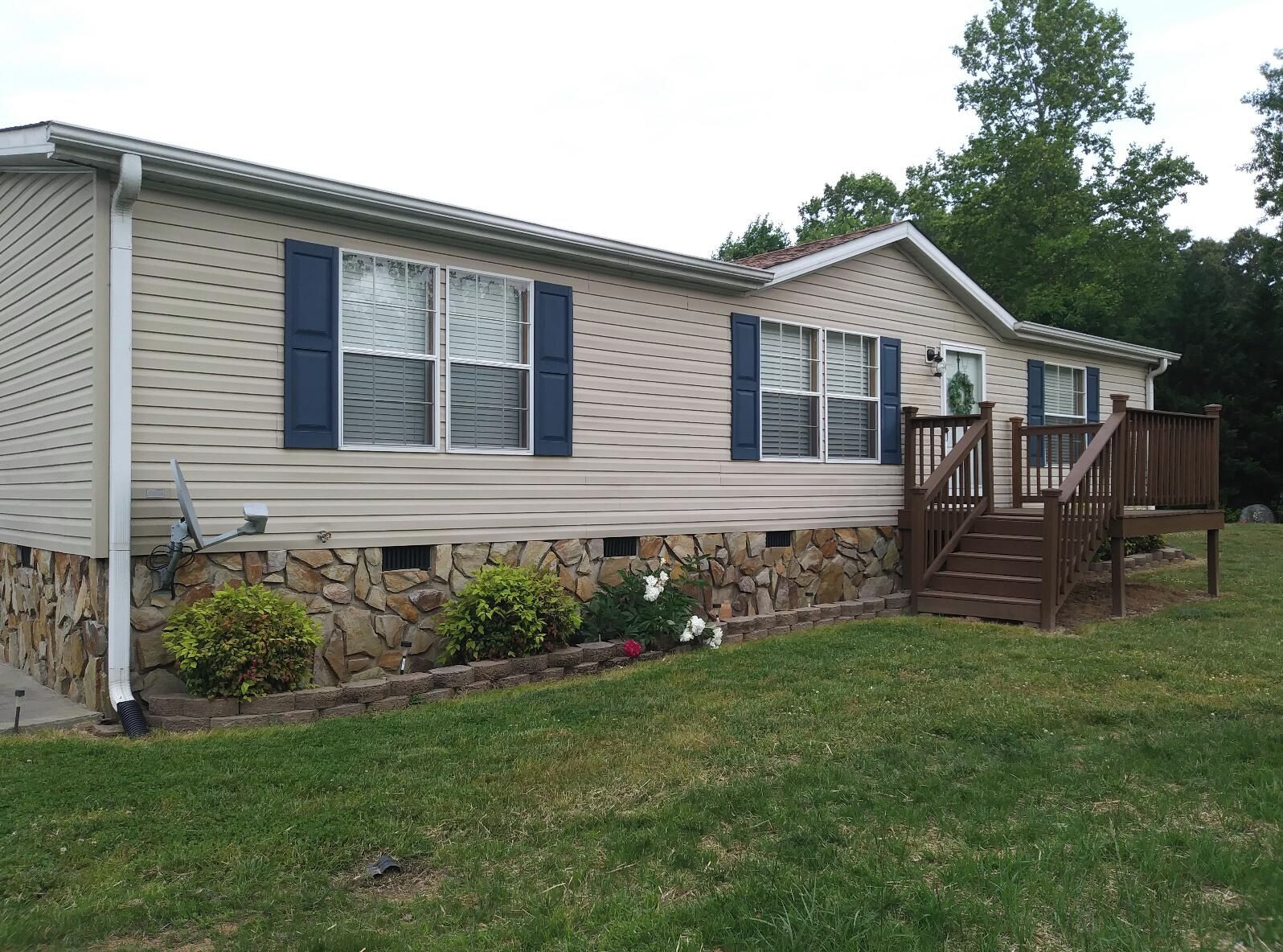 A single-story mobile home with a stone foundation, a front porch with a wooden staircase, and a small garden area.