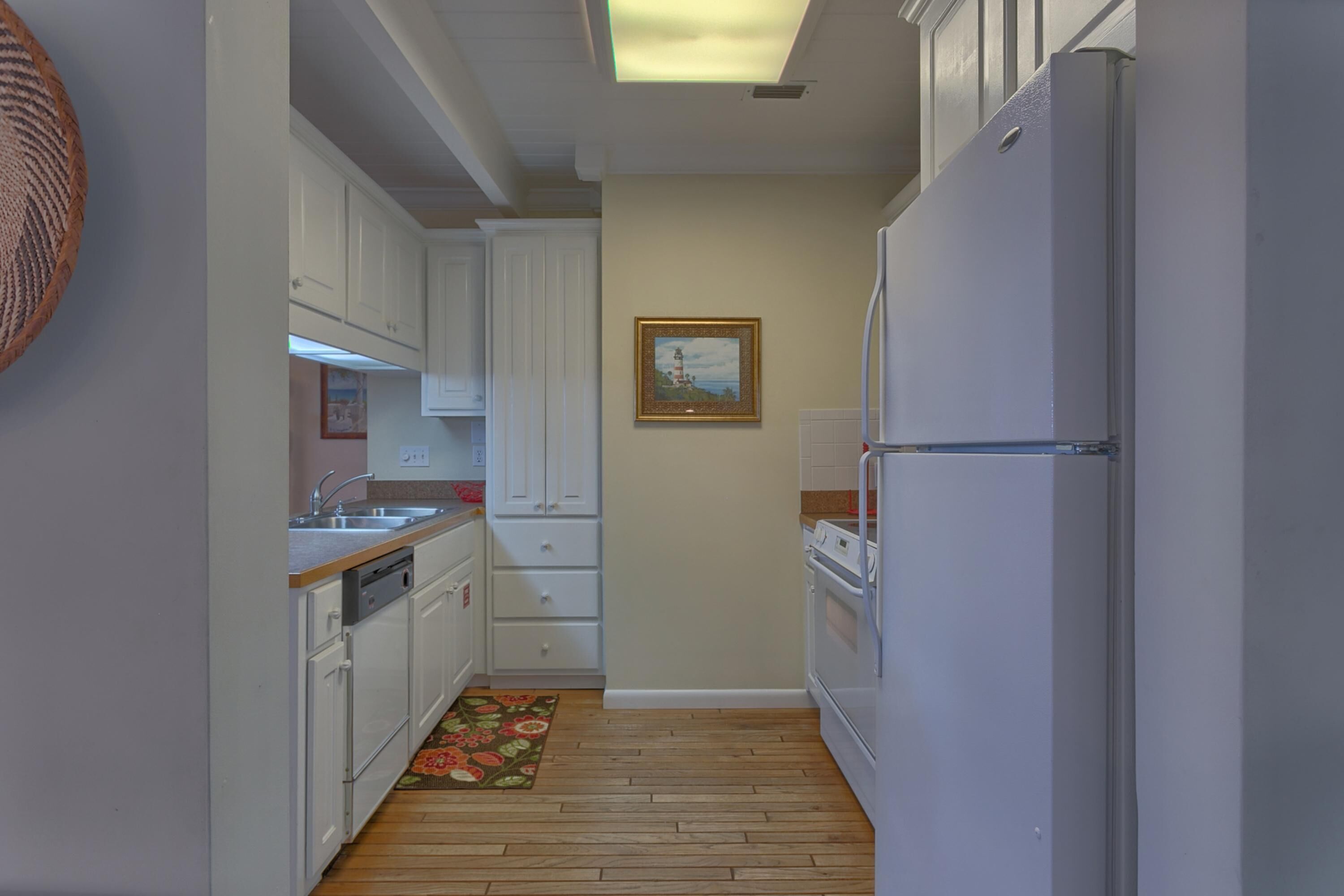 A narrow kitchen with white cabinets, a refrigerator, and a wooden floor.