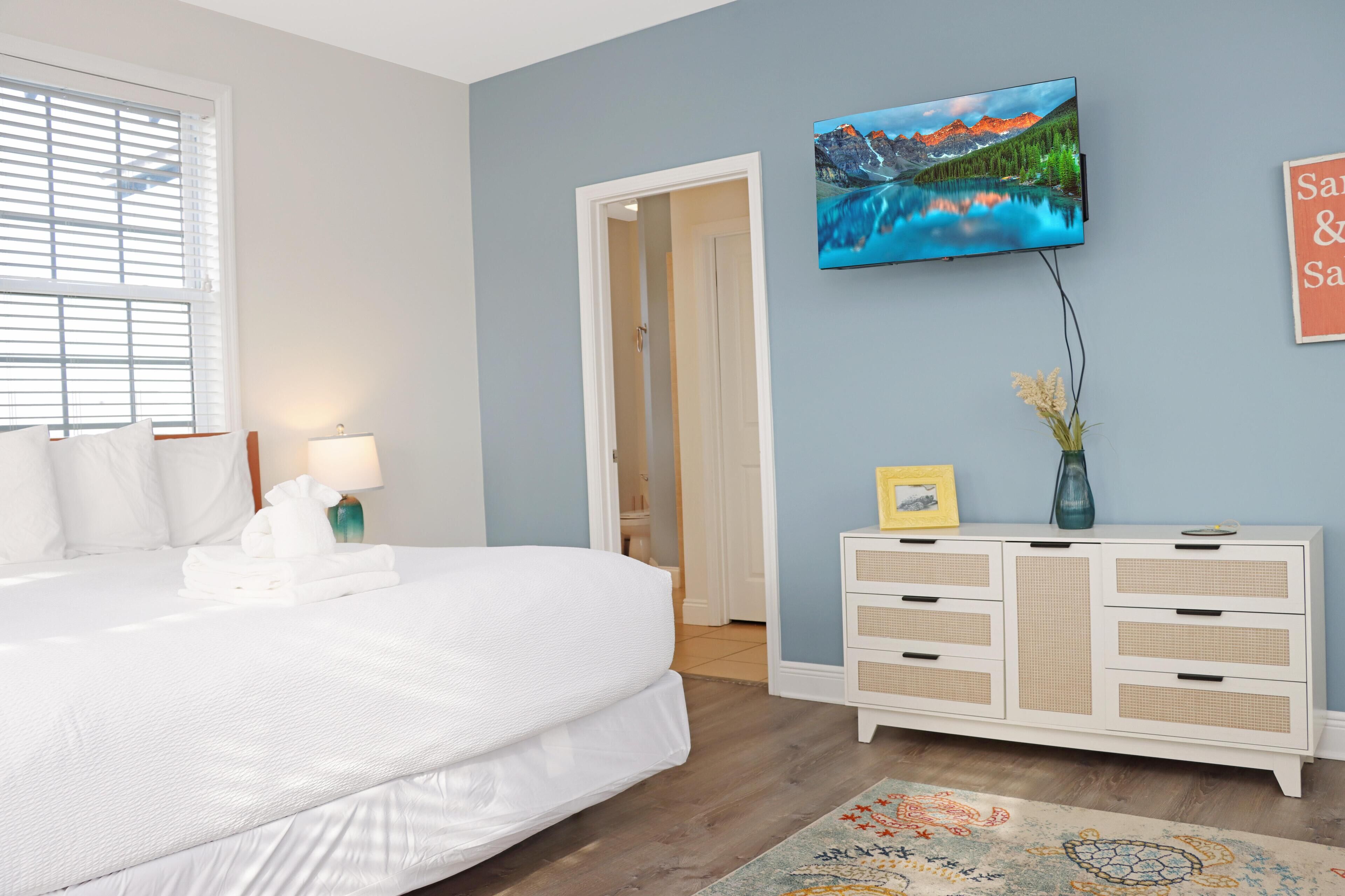 A bedroom with a large bed, a TV mounted on the wall, a dresser, and a door leading to another room.