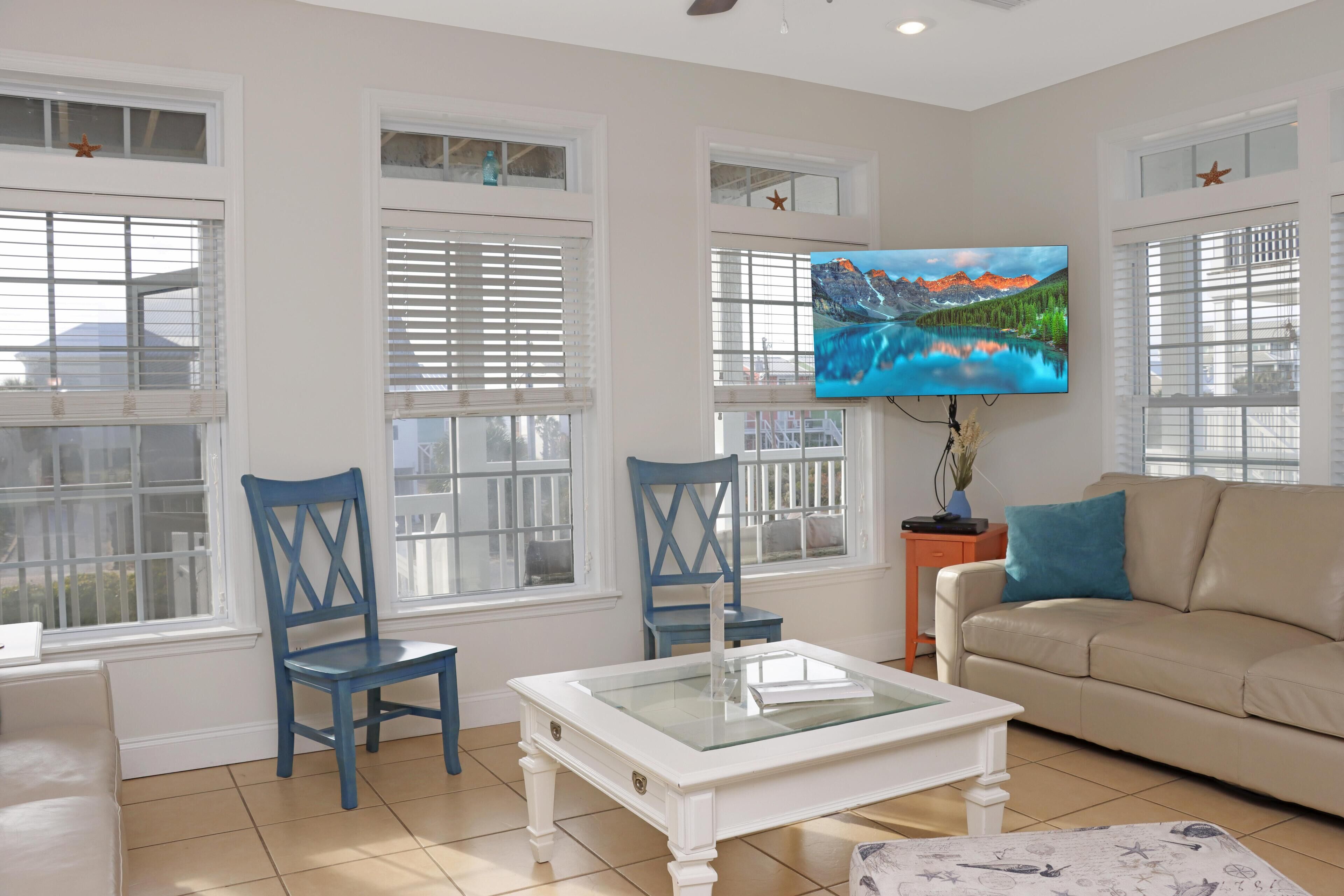 A living room with a beige sofa, two blue chairs, a white coffee table, and a TV mounted on the wall displaying a scenic landscape.