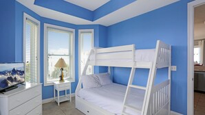 A bedroom with a bunk bed, a dresser, a nightstand with a lamp, and a window with blinds. - Cape Destiny (Port St. Joe)