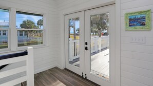 Una habitación con paredes blancas, suelo de madera, una banca, una puerta con paneles de vidrio y vistas a un área cercada con palmeras.