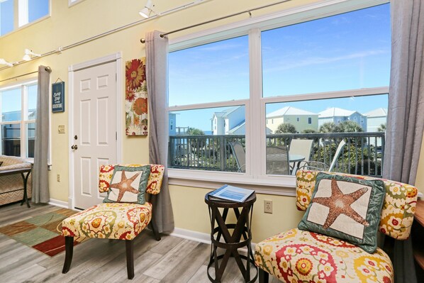 A room with two floral-patterned chairs, a small table with a book, and a large window with a view of houses and a balcony.