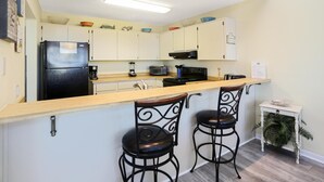 A compact kitchen with a refrigerator, microwave, toaster, and coffee maker. There are two bar stools and a small table with a bowl. - Coastal Sunshine (Port St. Joe)