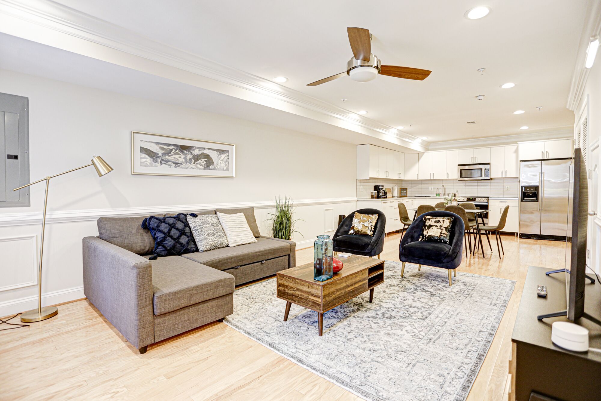 A modern living room with a sofa, armchairs, and a coffee table. There is a ceiling fan, recessed lighting, and a kitchen in the background.