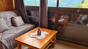 Ein gemütliches Zimmer mit Holzboden, einem braunen Sofa mit Kissen, einem kleinen Holztisch mit einer Laterne und einem großen Fenster mit Blick auf Grünflächen.