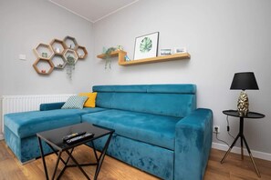 A living room with a blue sectional sofa, a black coffee table, a wall-mounted hexagonal shelf with decorative items, and a small round table with a lamp.