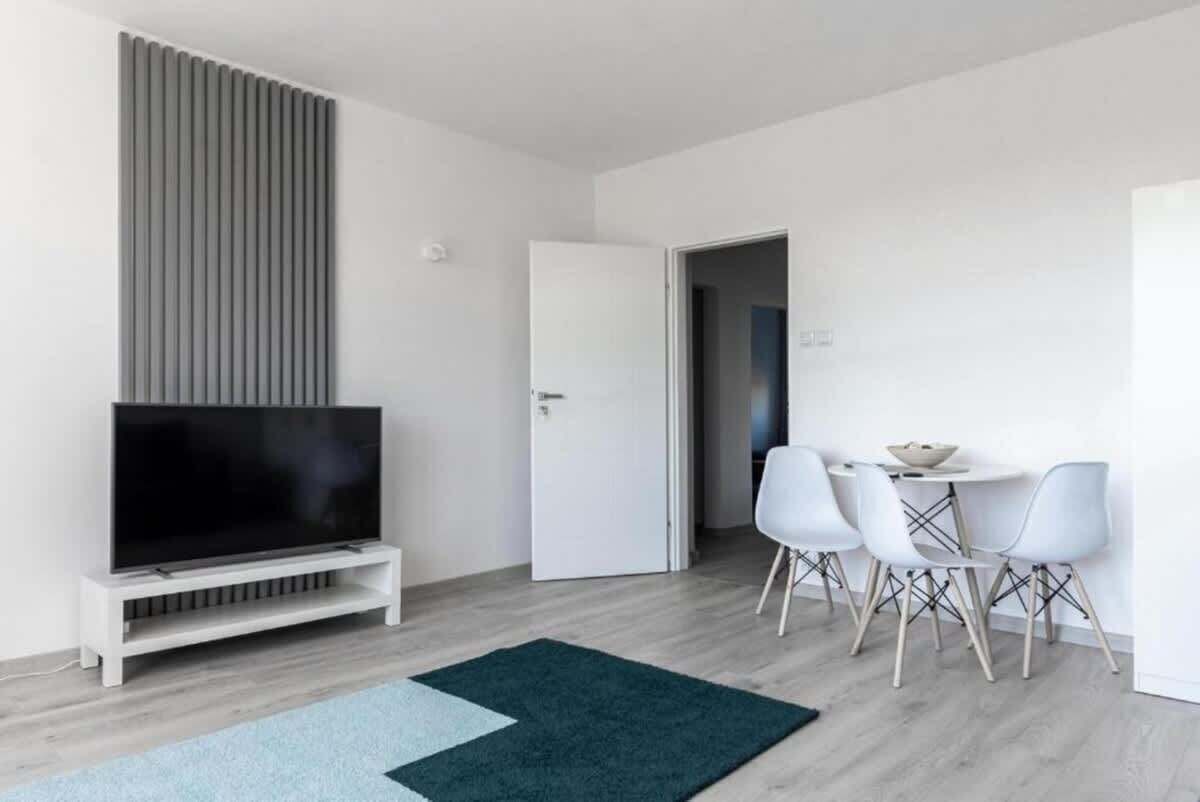 A modern living room with a flat-screen TV on a white stand, a dining area with a small table and four chairs, and a door leading to another room.