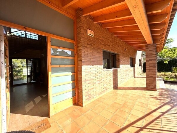An open patio area with a wooden ceiling and brick walls, a door leading to an interior space, and a tiled floor.