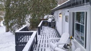 Une terrasse extérieure couverte, avec des chaises blanches et un barbecue, entourée de neige et d’arbres.