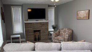 A living room with a grey sofa, a fireplace, a television, and a ceiling fan. - Holland Suites, Suite #1 (Williamstown)