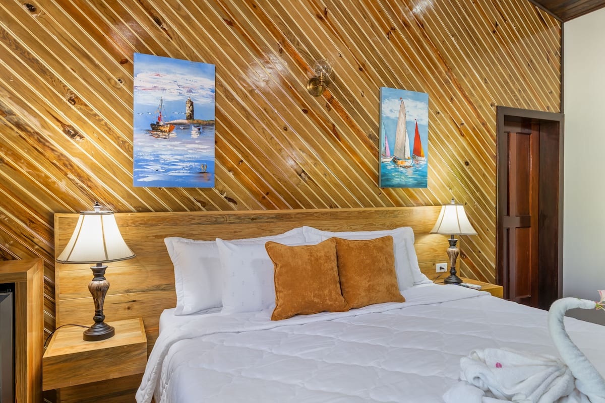 A hotel room with a wooden slat ceiling, a bed with white bedding, two lamps, and two paintings of sailboats.