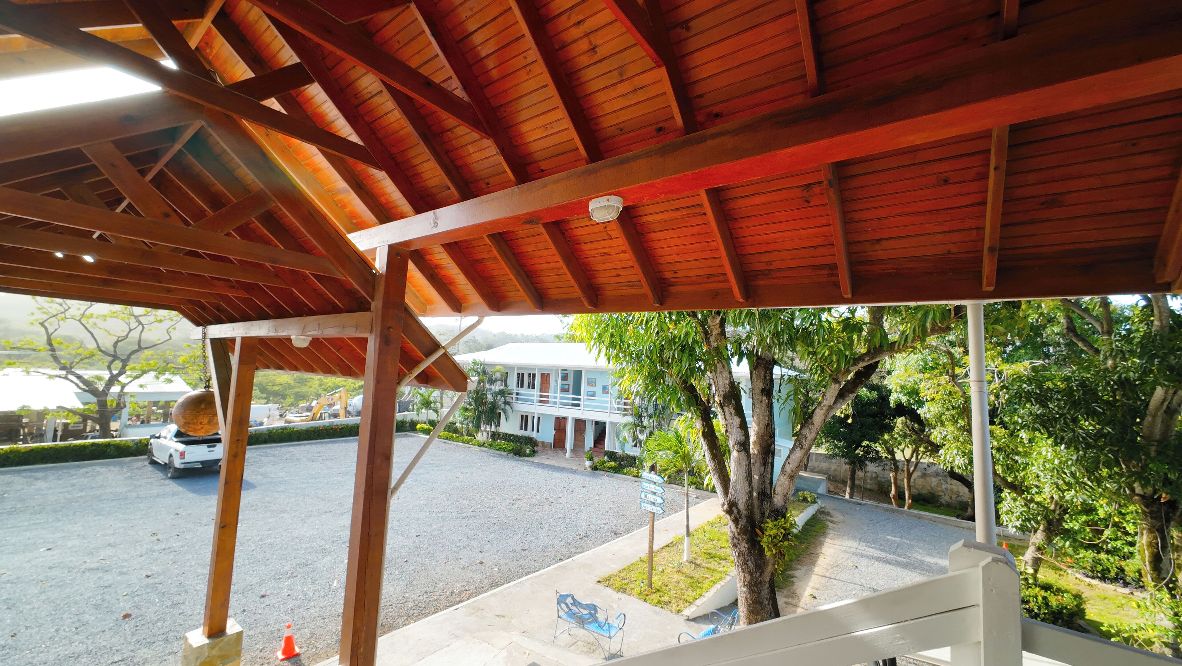 Un espacio al aire libre techado con estructura de madera, un camino pavimentado y un edificio al fondo.