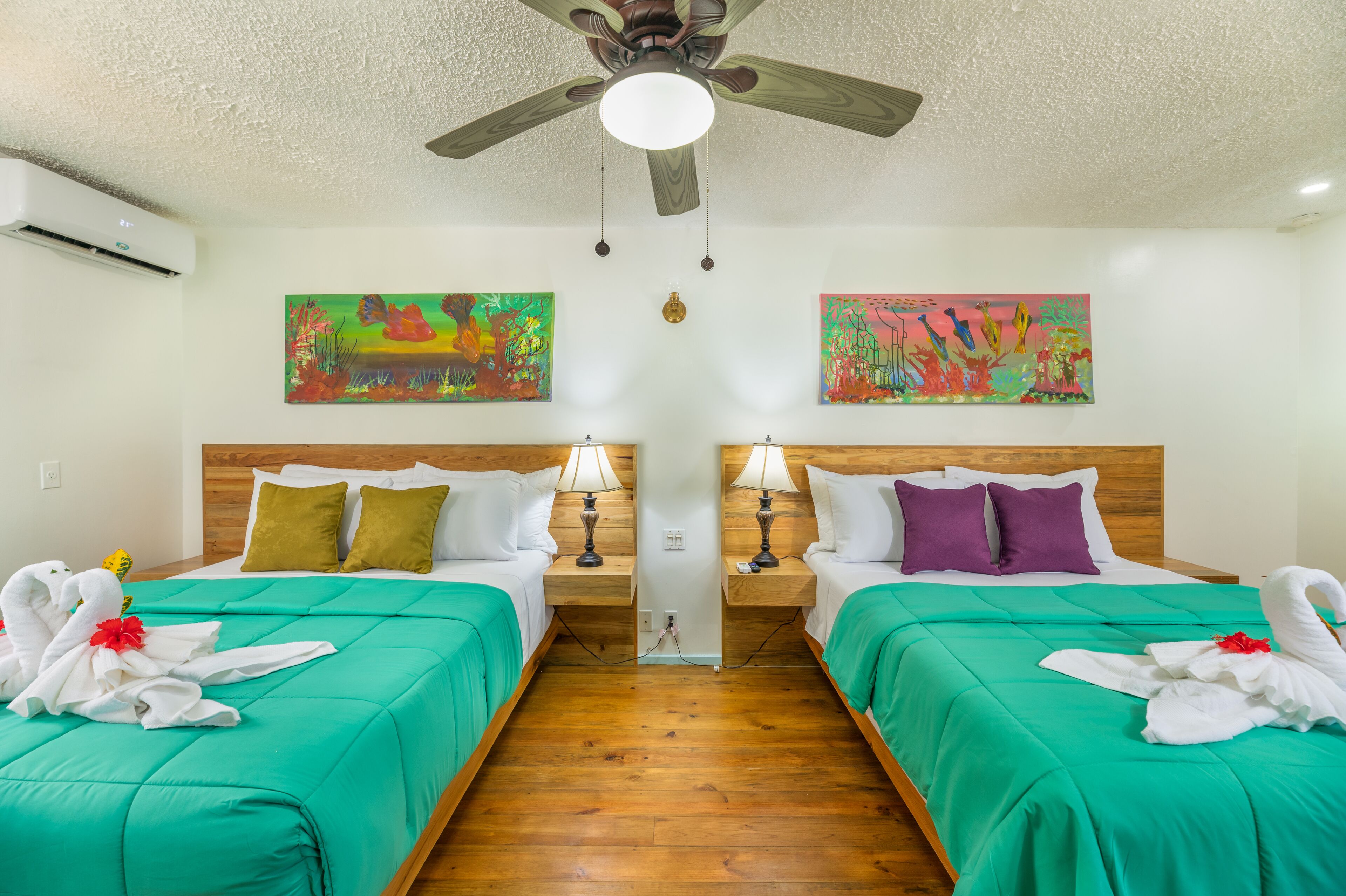 Una habitación de hotel con dos camas, ropa de cama verde, toallas blancas con rosas rojas, cabeceros de madera, lámparas de noche y dos cuadros coloridos en las paredes.