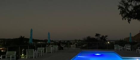 Piscina iluminada con luz azul, rodeada de sillas y sombrillas blancas.