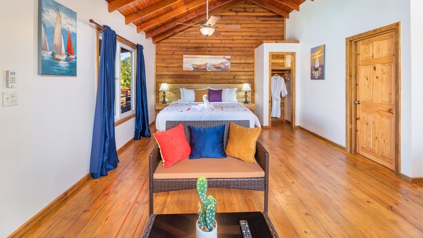 A spacious bedroom with a wooden ceiling, a bed with multiple pillows, a wicker sofa, and a cactus on a table.