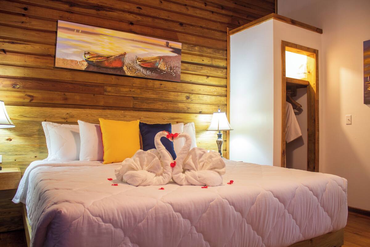 A bed with a white quilt and two swans made of towels with red hearts. There are pillows in yellow and white, and a painting of a boat on the wall.