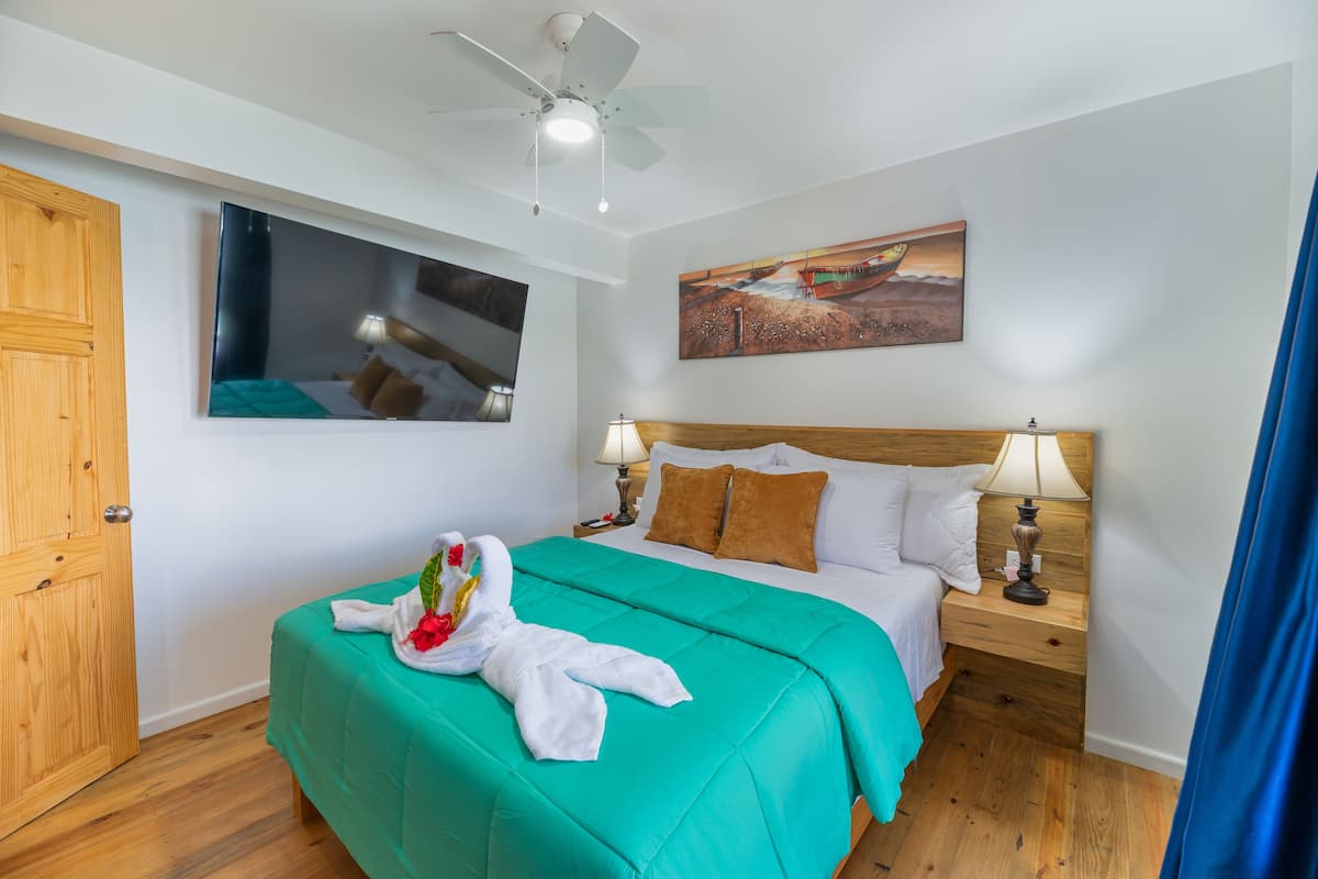 A hotel room with a bed, wooden headboard, two bedside lamps, a ceiling fan, a painting, and a wooden door.