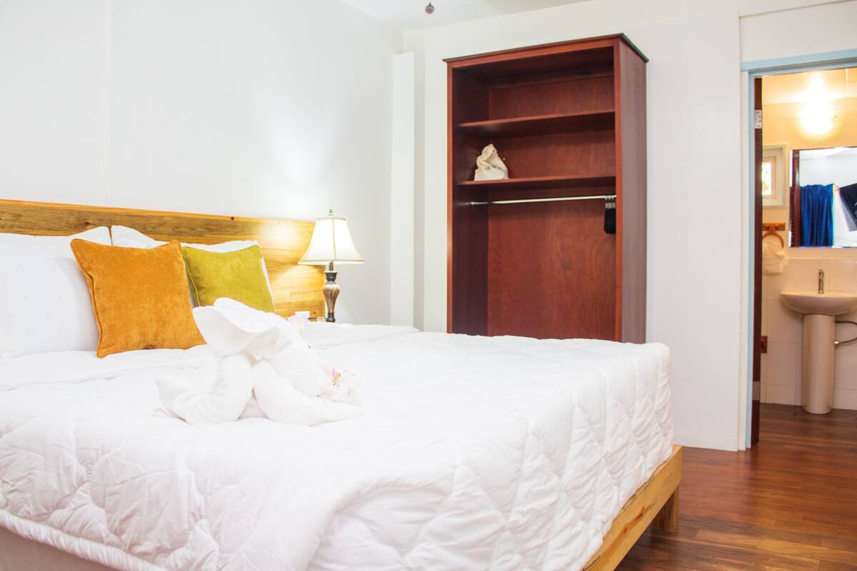 A hotel room with a large bed, a wooden wardrobe, a nightstand with a lamp, and a bathroom visible through an open door.