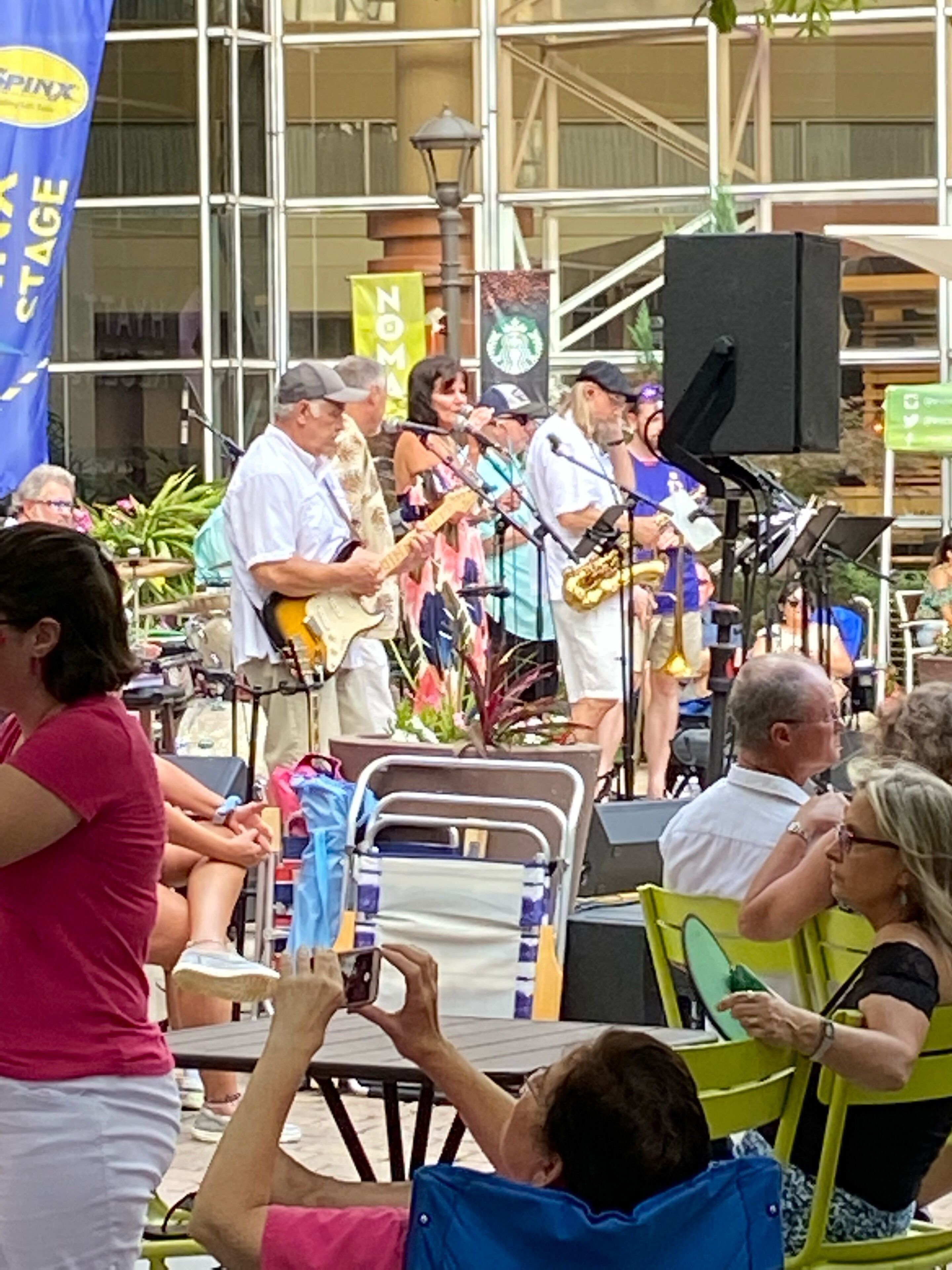 A band performing outdoors with a stage and audience seated on chairs.