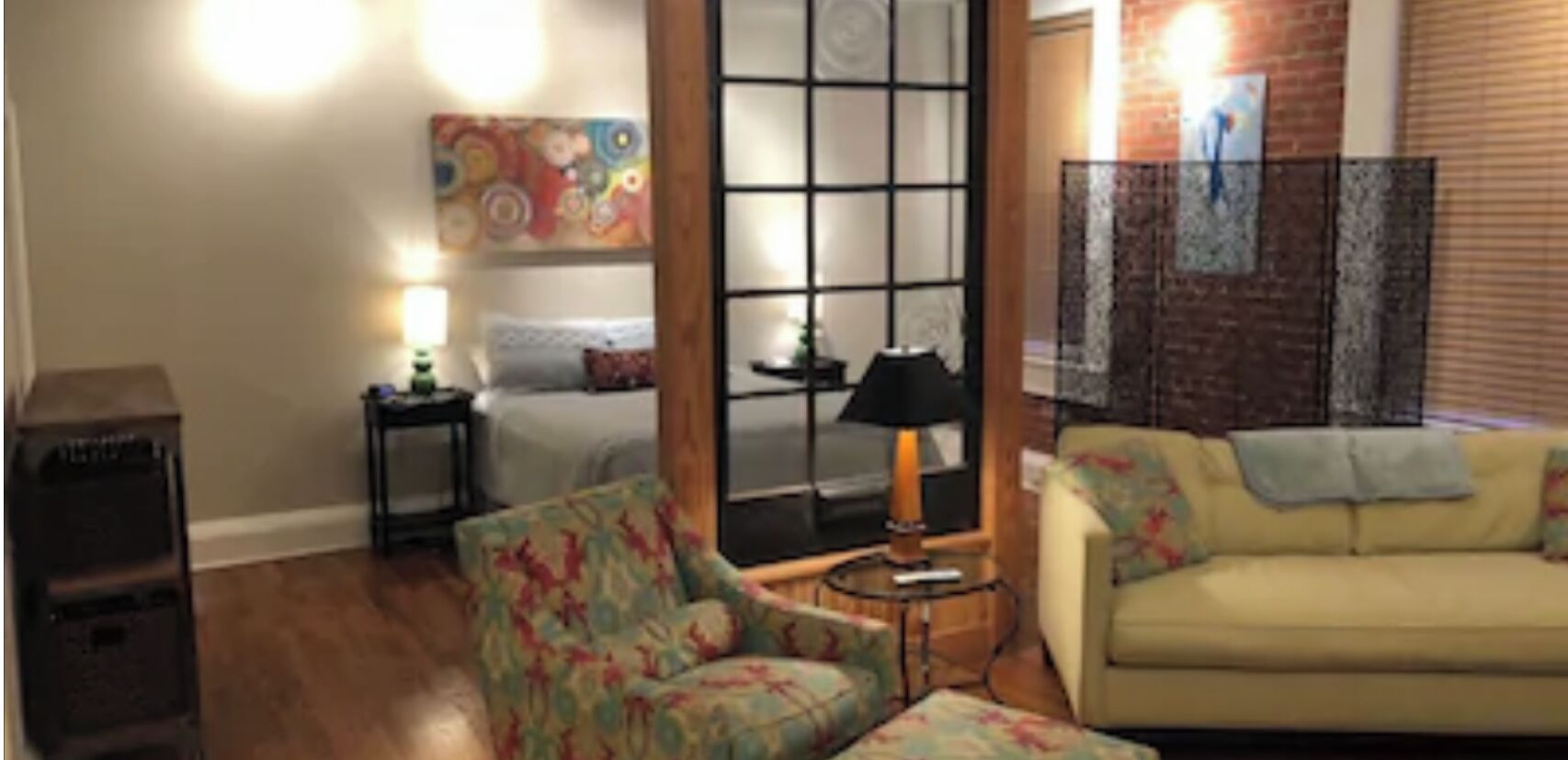 A living room with a sofa, armchair, and a small table with a lamp. There is a bed with a headboard and a painting on the wall.
